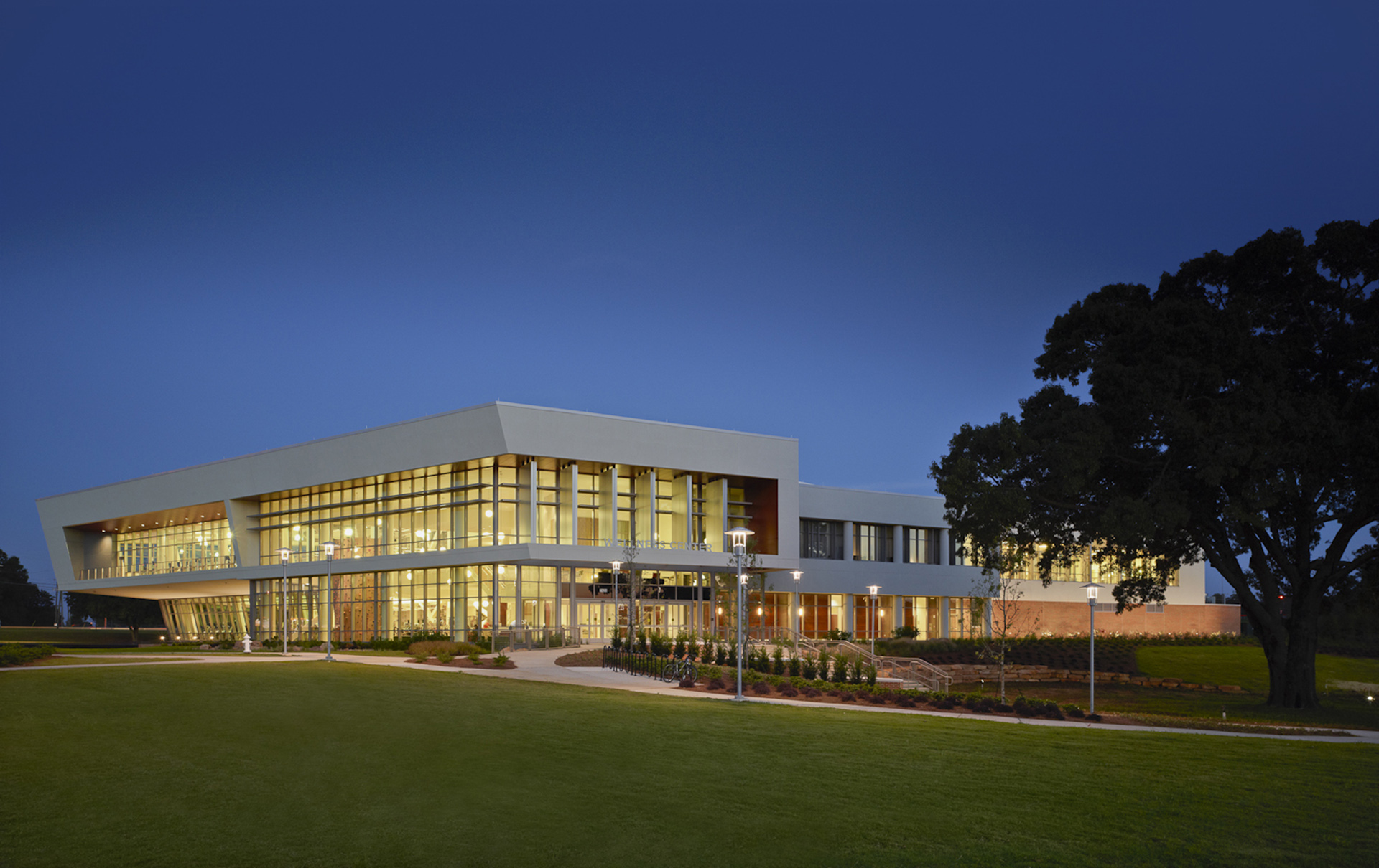 A long, low glass-fronted building at dusk, lit from inside, with a live oak on the right